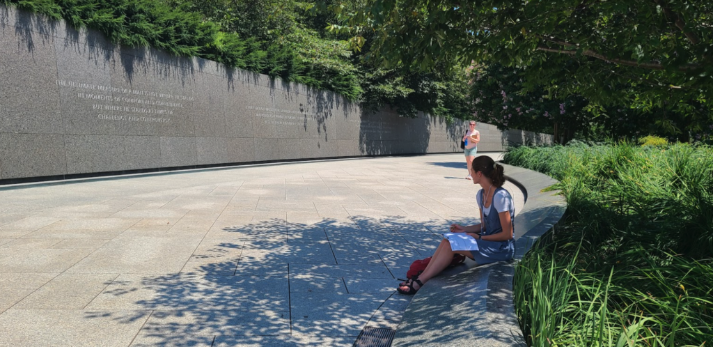 A woman sits under a shade tree on the right, facing the Vietnam War Memorial wall.