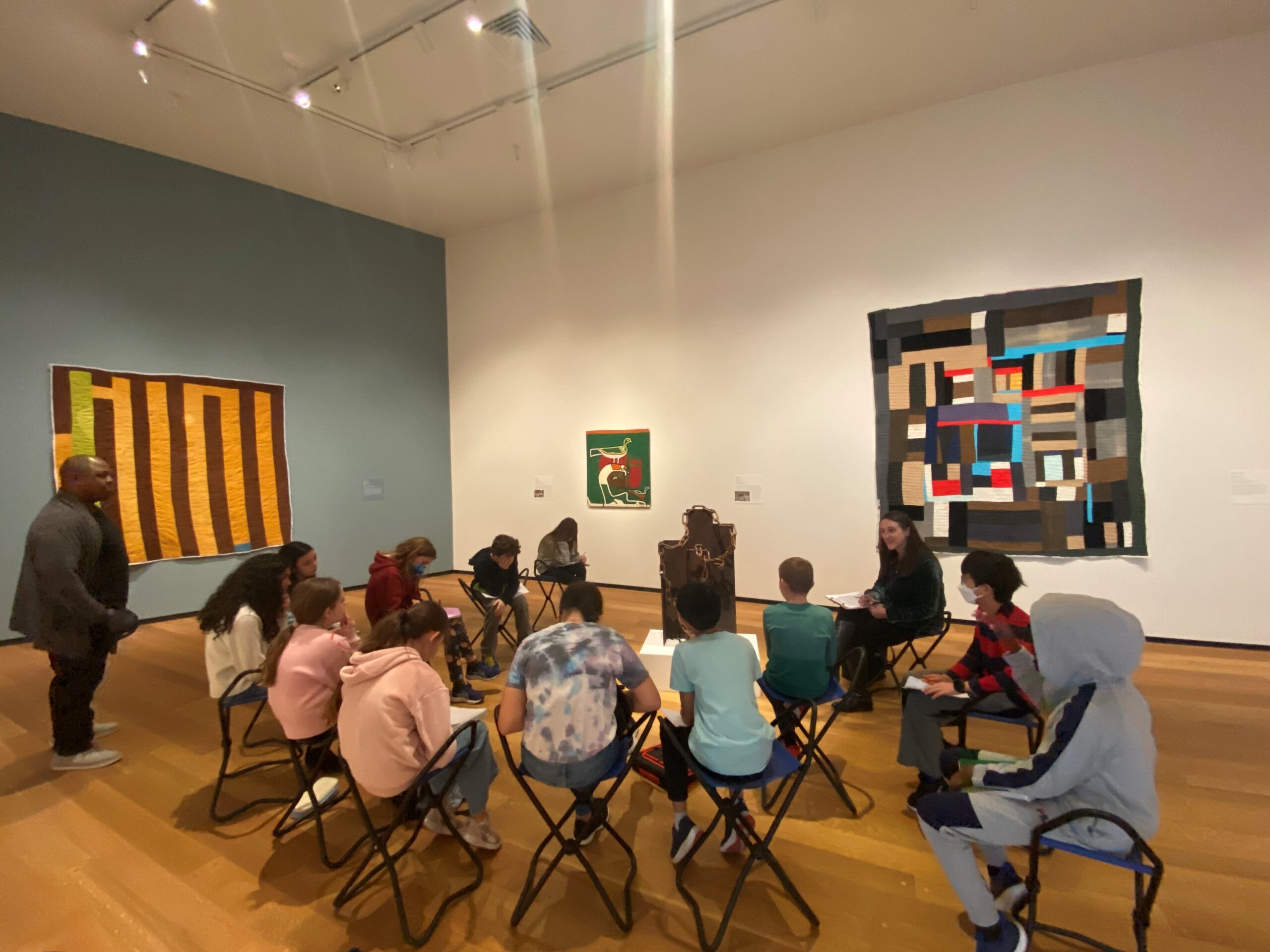 A group of middle-school students sit in stools around a museum educator in an art gallery.