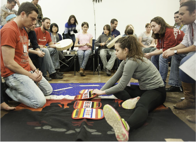 A group of teens circle around a girl and a boy playing the Moccasin game. The girl reaches her hand under one of the colorful patterned "moccasins" while the boy waits.