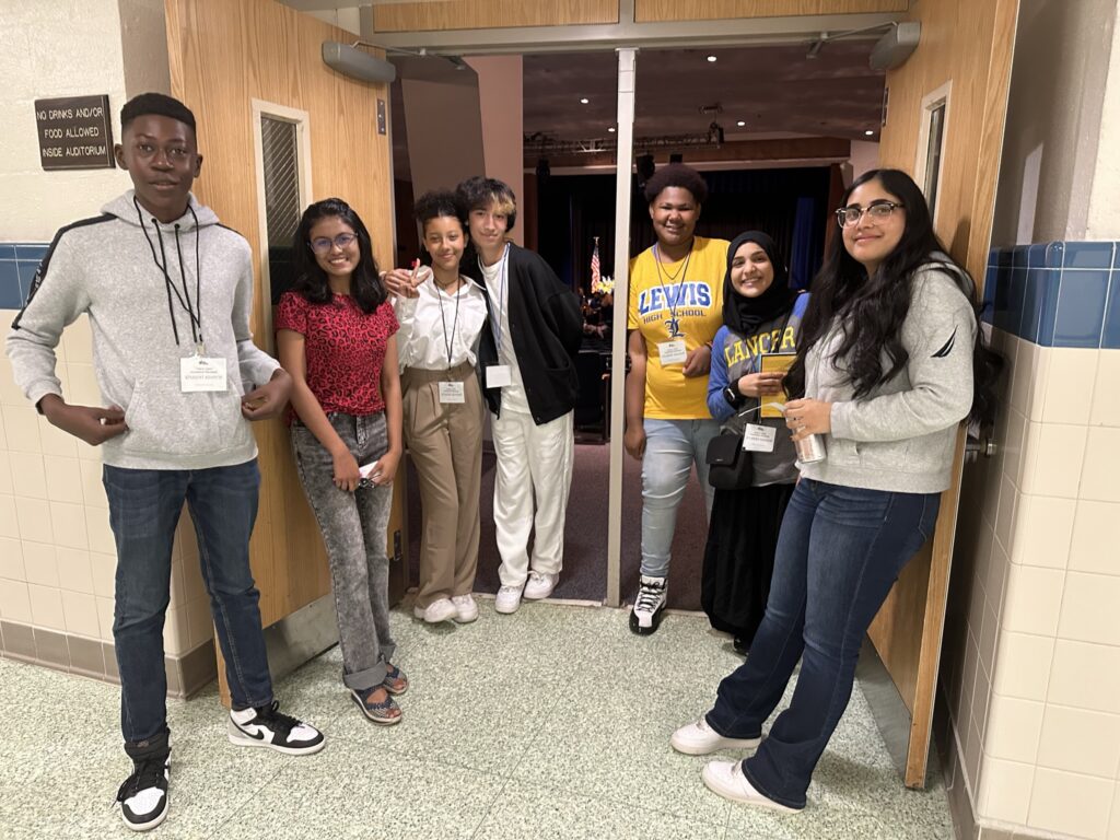 A racially diverse group of teenagers pose, smiling, in the doorway to an auditorium.
