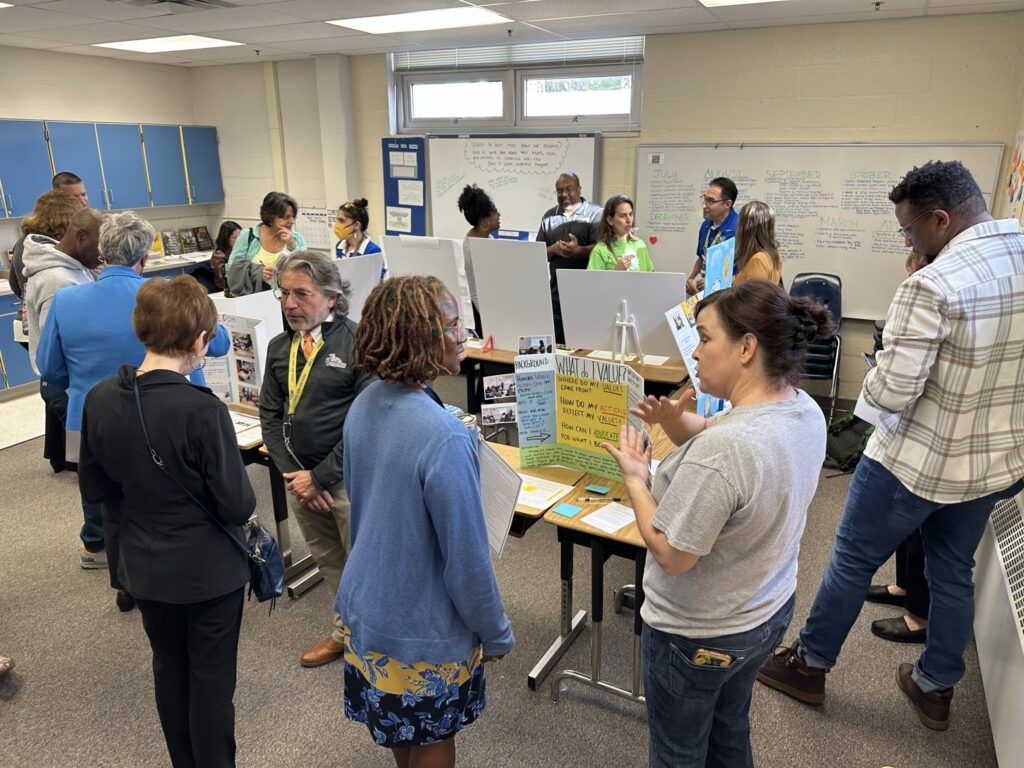 Students and teachers stand around a classroom exhibition, engaged in deep conversation with each other.