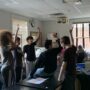 Students raise their hands eagerly while standing around a table in their classroom.