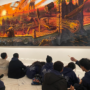 Ms. Fisher's 8th grade students sit on the gallery floor, looking up and gesturing towards the large painting on the wall (Manifest Destiny).