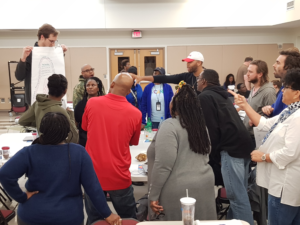 A large group of racially diverse teachers are standing around a table and looking at one female teacher who is standing on a chair and holding up a large sheet of paper. One male teacher is pointing towards the paper.