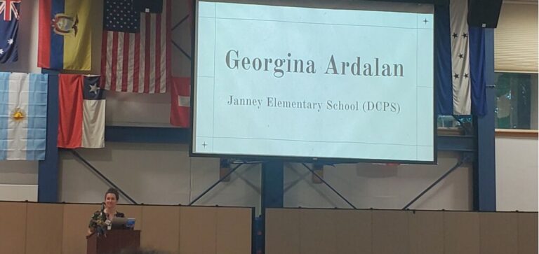 Georgina presents during a WISSIT 2023 plenary. She stands at a podium on a stage with a screen that shows her name behind her.