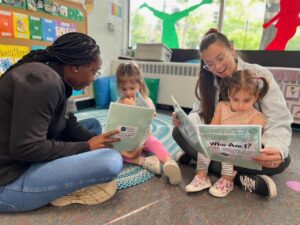 Two teachers sit on the ground and read with two young students.